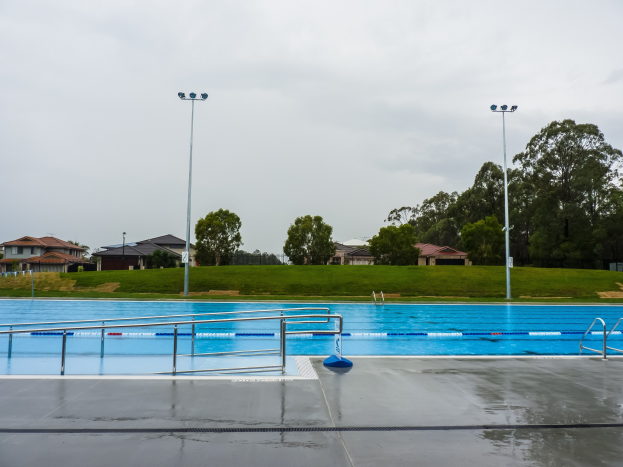 Gro├čer Swimmingpool in einem Park, umgeben von Z├Ąunen, Pfosten, Lichtern und B├Ąumen, mit H├Ąusern und einem klaren blauen Himmel im Hintergrund.
