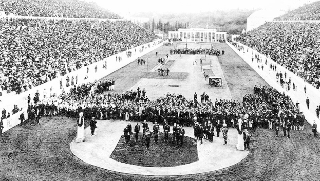 Schwarzes und weißes Foto einer großen Menge in einem Stadion während der Eröffnungszeremonie der Olympischen Spiele 1896, mit Menschen auf den Rängen und anderen auf dem Boden sitzend oder stehend.