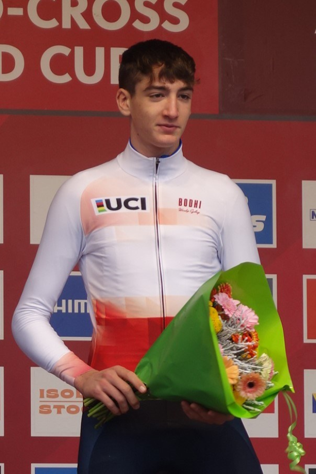 Ein Mann in einem Anzug steht auf einem Podium und hält einen Blumenstrauß, feiernd mit einem selbstbewussten und stolzen Ausdruck.
