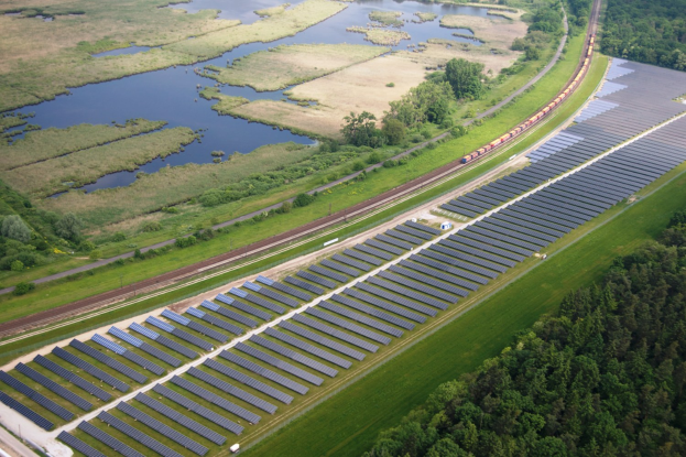 Luftaufnahme eines großen Solarparks mit Solarpanelen in einem Feld, umgeben von Bäumen, Gras, Pflanzen und Wasser, mit einem Zug auf einer nahen Eisenbahnstrecke.