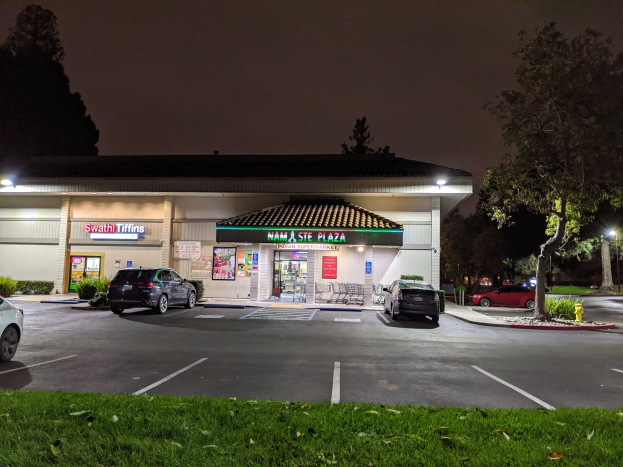 Ein nächtlicher Parkplatz mit geparkten Autos, beleuchtet von Lampen, umgeben von Gras, Pflanzen, Bäumen und einem Feuerhydranten, mit einem Geschäft im Vordergrund und dem Himmel im Hintergrund.