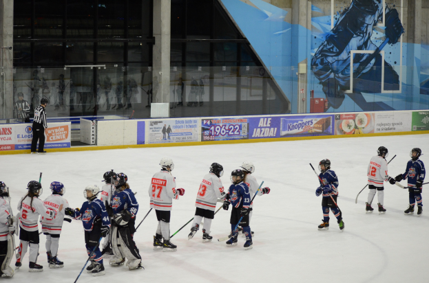 Eine Gruppe junger Menschen, die Eis hockey auf einer Indoor-Eisbahn spielen, tragen Helme und halten Stöcke, mit einer Person, die links steht und Bannern, Säulen und Glaswänden im Hintergrund.