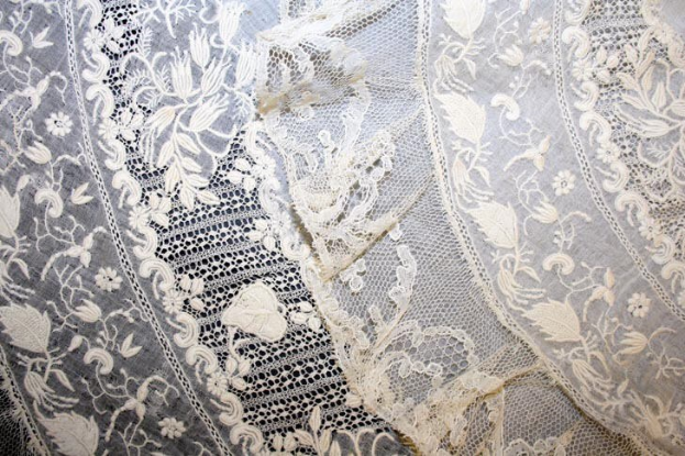 Nahaufnahme eines weißen Hochzeitskleids mit feiner Spitzenverzierung mit zarten, verschiedenen Mustern auf einem leichten, luftigen Stoff.