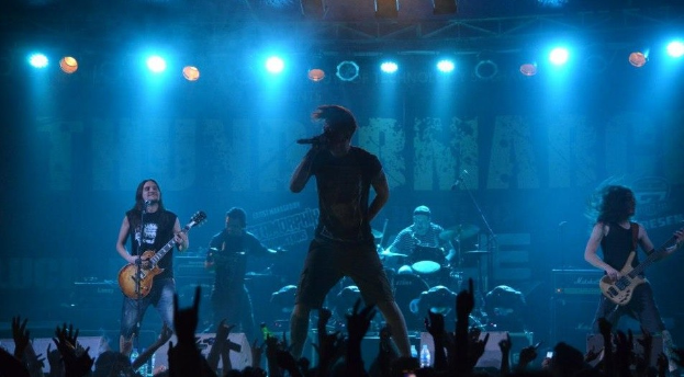 Eine Gruppe von Musikern, die auf einer hell erleuchteten Bühne mit Gitarren, einem Mikrofon und Trommeln spielen, während die Zuschauer im Vordergrund die Hände heben.