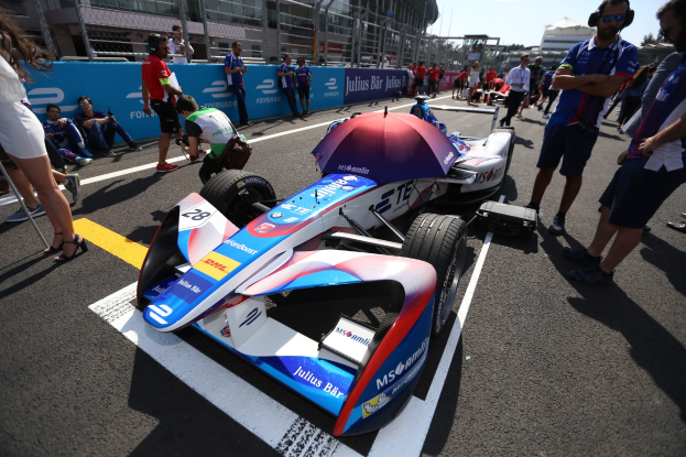 Formula E-Auto mit weißer, blauer, roter und schwarzer Lackierung auf einer Rennstrecke geparkt, umgeben von Menschen, mit einem Schirm im Vordergrund und sichtbarer Rennstrecken-Infrastruktur im Hintergrund unter einem klaren blauen Himmel.