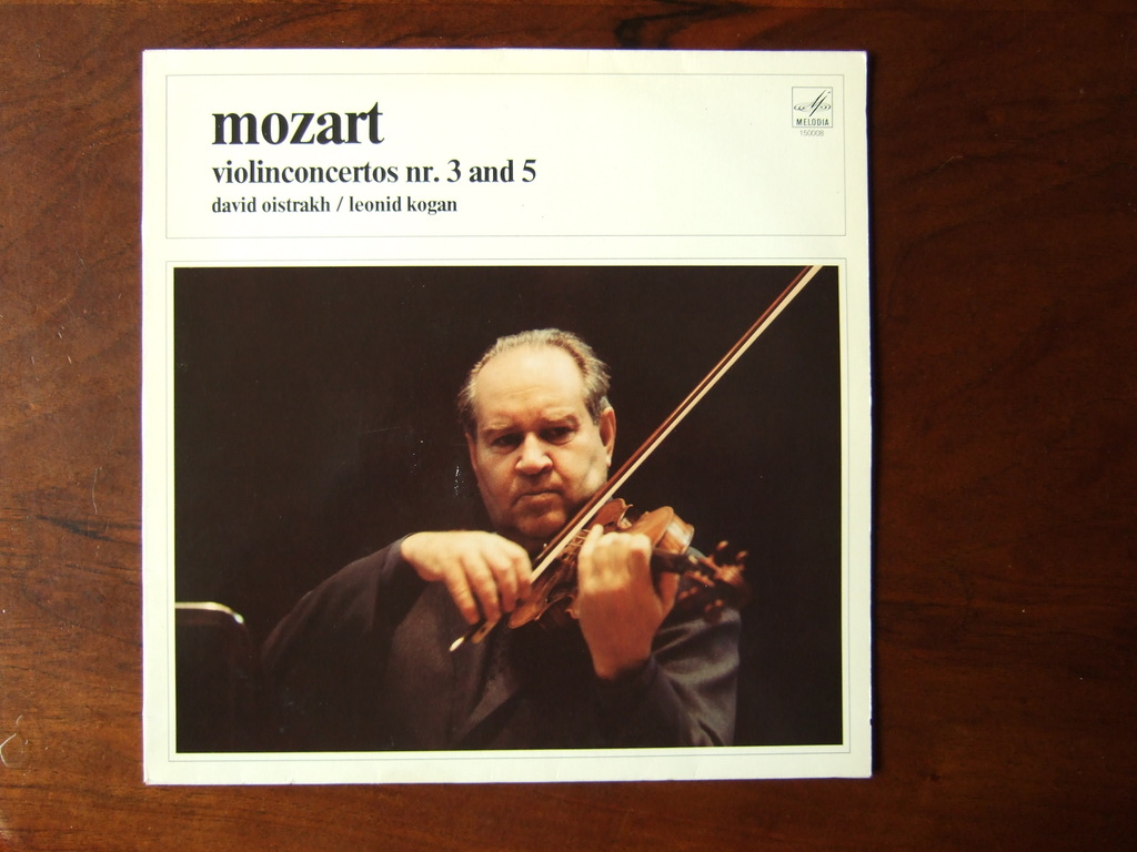 Ein Mann in einem schwarzen Mantel spielt Geige auf einem Holztisch, mit schwarzem Hintergrund und einer kleinen Inschrift oben auf dem Bild.