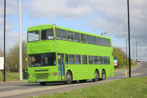 Grüner Doppeldeckerbus auf einer Straße mit Gras, Strommasten, Schildern, Bäumen und anderen Fahrzeugen im Hintergrund unter einem bewölkten Himmel.