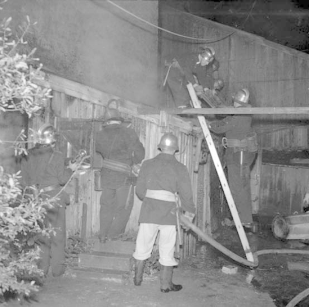 Feuerwehrleute in Helmen stehen auf Treppenstufen, einer auf einer Leiter und ein anderer hält ein Rohr, mit einem Baum links und einer Wand im Hintergrund, in Schwarz-Weiß.