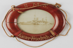Rotes Rettungsfloß mit Text "USS Kaiser Friedrich III" und Bild eines Schlachtschiffs, an einer weißen Oberfläche mit Seilen befestigt.