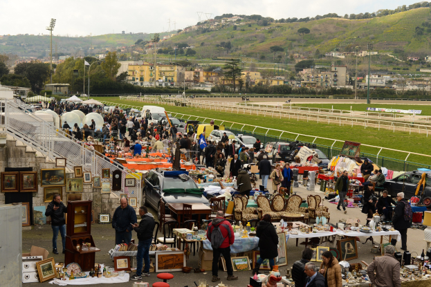 Eine große Gruppe von Menschen auf einem Flohmarkt mit Tischen, auf denen Gegenstände wie Foto rahmen und Stühle ausgelegt sind, daneben geparkte Fahrzeuge, Geländer, Stufen, Bäume, Gebäude, Laternenpfähle, Hügel und ein bewölkter Himmel im Hintergrund.