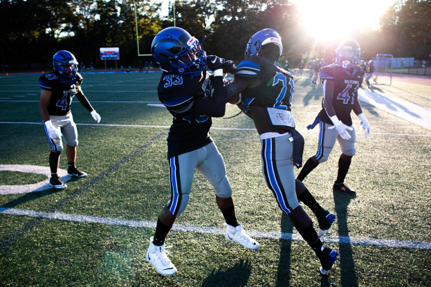 Eine Gruppe junger Männer, die Fußball auf einem Feld mit Bäumen und einem klaren blauen Himmel spielen, tragen Helme und Schuhe.