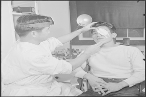 Ein Schwarz-Weiß-Foto eines Mannes, der von einem stehenden Arzt untersucht wird, mit verschiedenen Gegenständen auf einem Tisch und einem Fenster mit einem Vorhang im Hintergrund.