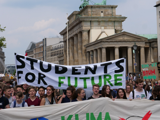 Gruppe von Schülern marschiert in Berlin mit einer bunten 'Students for Future'-Schriftzug vor dem Hintergrund von Gebäuden, Bäumen und Himmel.