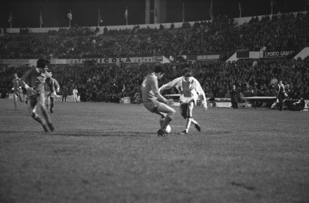 Schwarzes und weißes Foto von Männern, die auf einem Feld Fußball spielen, mit Zuschauern auf den Tribünen und Fahnen im Hintergrund.