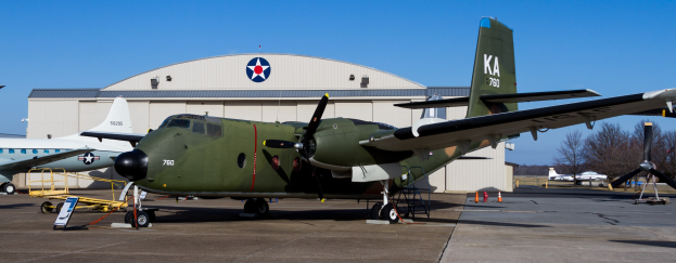 Grüner Flieger auf einem Flughafenvorfeld neben einem Gebäude, umgeben von Absperrgittern und Schildern, mit Bäumen und einem klaren blauen Himmel im Hintergrund.