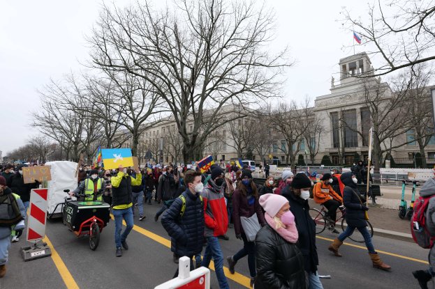 Eine große Gruppe von Menschen marschiert auf einer Demonstration in Washington, D.C. und hält Schilder und fährt Fahrräder, mit Bäumen und einem klaren blauen Himmel im Hintergrund.