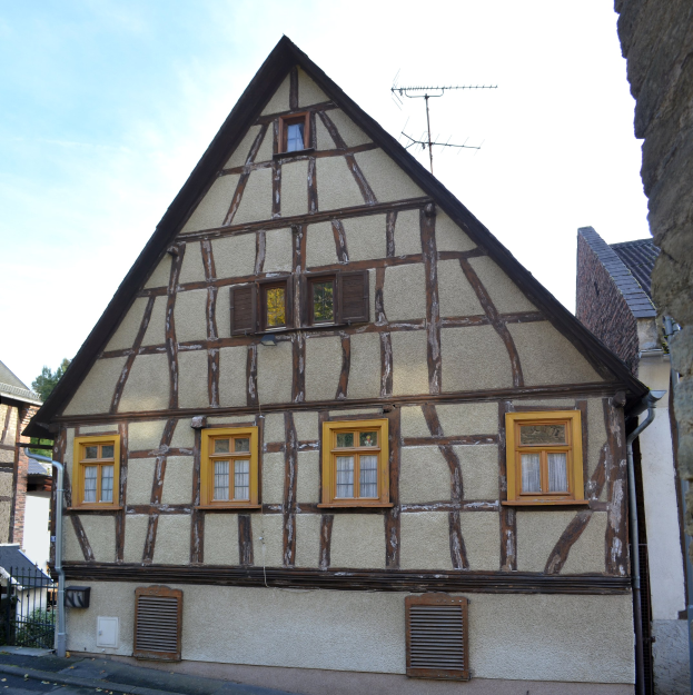 Alter Fachwerkbau mit mehreren Fenstern, umgeben von anderen Gebäuden, Bäumen und einem klaren blauen Himmel.