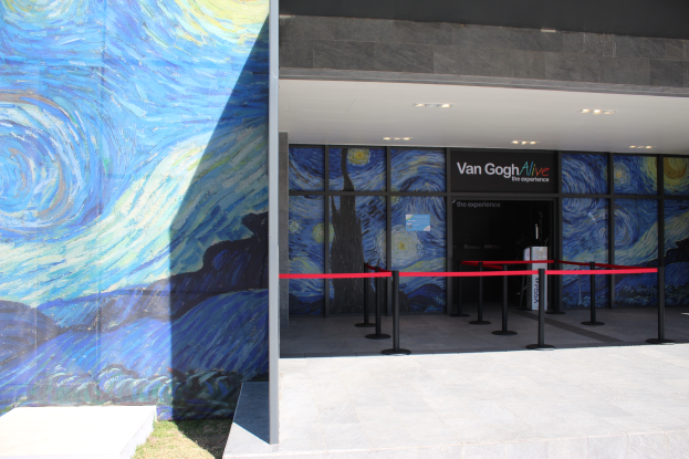 Der Eingang eines Gebäudes zeigt ein großes Wandbild, ein Schild mit der Aufschrift "Van Gogh Alive", Absperrpoller mit Bändern, Gras auf dem Boden und Lampen an der Decke.