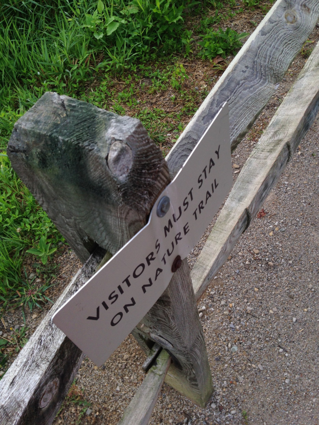 Holzzaun mit einem Schild, auf dem "Besucher sollten den Naturweg nicht verlassen", umgeben von grünem Gras und kleinen Steinen.