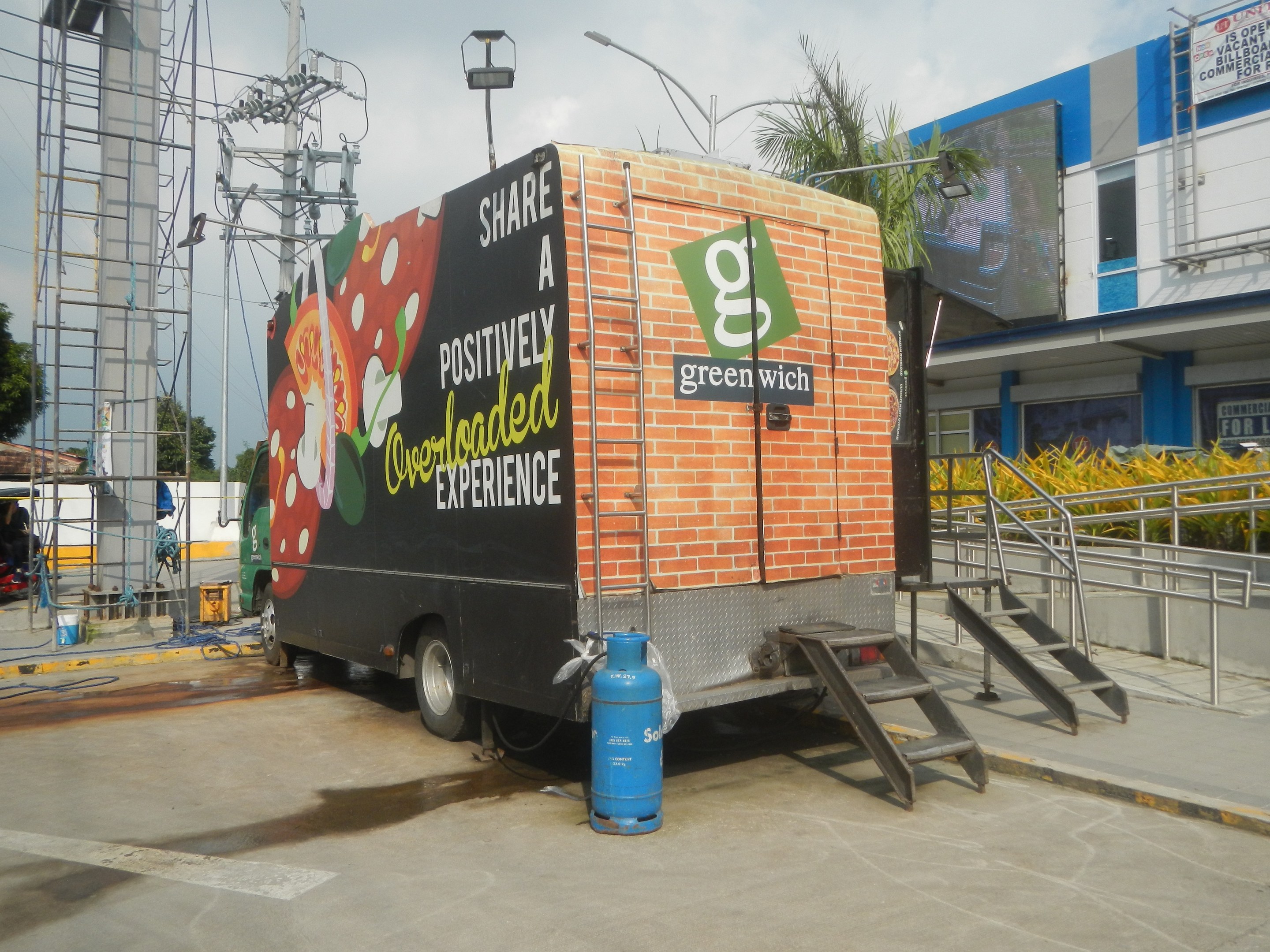 Ein Lkw vor einem Gebäude mit einer Tafel, auf der "Teilen Sie ein positiv überladenes Erlebnis" steht, mit Stufen, einem Zylinder auf dem Boden, Geländern, Strommasten mit Drähten, Laternenmasten, Bäumen und einer bewölkten Himmel.