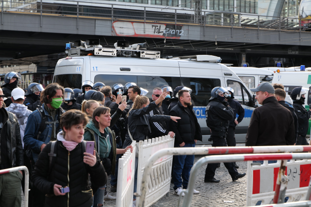 Eine Gruppe von Menschen steht vor Polizeifahrzeugen, einige tragen Helme und halten Telefone, mit Absperrungen im Vordergrund und einer Brücke und Gebäuden im Hintergrund in Berlin, wo die Polizei Protestierende auseinander treibt.