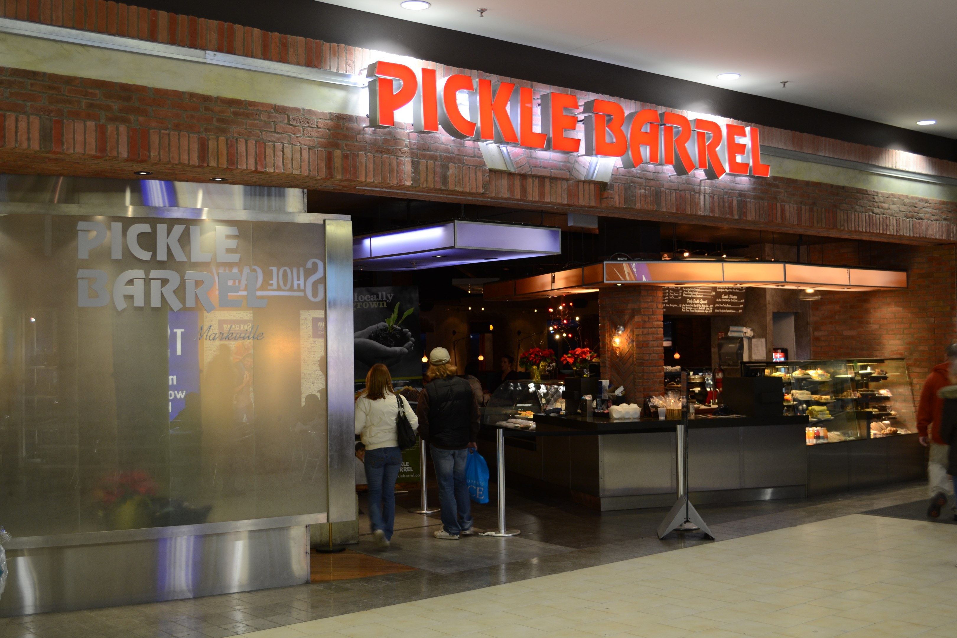 Ein Pickle-Bar-Restaurant in einem Einkaufszentrum mit Menschen, einem Schild, einer Auslage, Pflanzen, einem Metallpfosten und Deckenleuchten.
