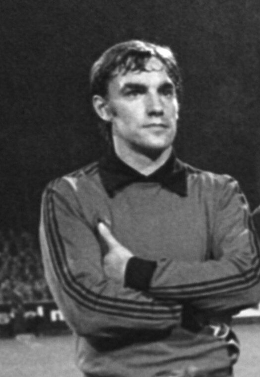 Ein Schwarz-Weiß-Foto eines älteren Mannes, der mit verschränkten Armen auf einem Fußballfeld steht, umgeben von einer Gruppe von Menschen im Hintergrund.