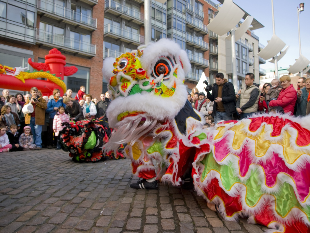 Ein lebendiges chinesisches Neujahrsfest in Amsterdam mit einer Drachenvorführung vor einer Zuschauermenge, einige halten Kameras, vor einer Kulisse aus Gebäuden, Laternenmästen und einem klaren blauen Himmel.