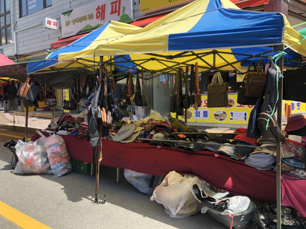 Ein Markt mit Zelten, die Taschen, Hüte und andere Artikel präsentieren, neben Gebäuden mit Fenstern und Texttafeln.