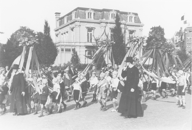 Eine Gruppe von Menschen in Schwarz und Weiß, die eine Straße entlanggehen und Fahnen halten, mit einem Maibaum im Hintergrund.