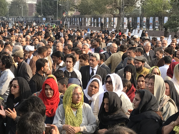 Große Menschenmenge protestiert vor einem Gebäude in Lahore mit Bäumen, Pfählen und Fahrzeugen im Hintergrund.