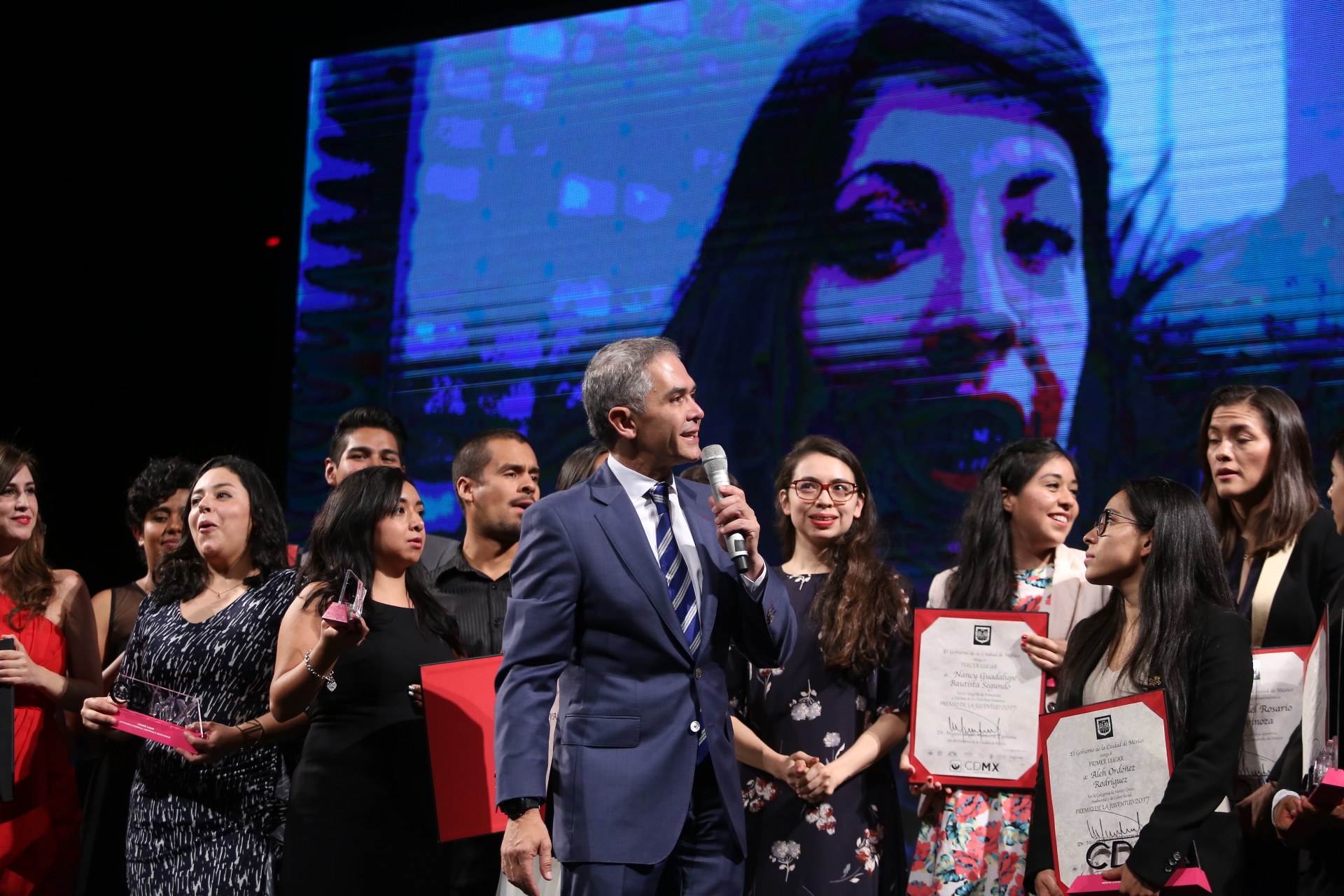 Ein Mann im Anzug und Krawatte spricht auf einer Bühne, während das Publikum Zertifikate hält, mit einem Bild einer Frau im Hintergrund.