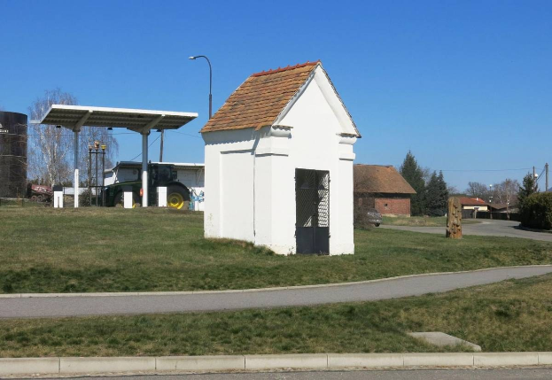 Ein kleines weißes Gebäude mit einem roten Dach, das als erste Tankstelle in Deutschland identifiziert wurde, steht an der Seite einer Straße umgeben von Gras, Bäumen, Polen, Laternen und Häusern mit einem klaren blauen Himmel im Hintergrund.