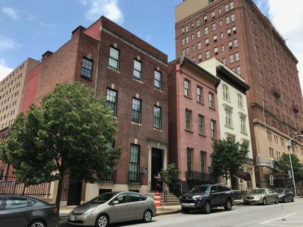 Eine von parkenden Autos gesäumte Straße mit Gebäuden, Bäumen, Laternen, Verkehrskegeln und einem Geländer unter einem bewölkten Himmel, die eines der teuersten Apartments in NYC zeigt.