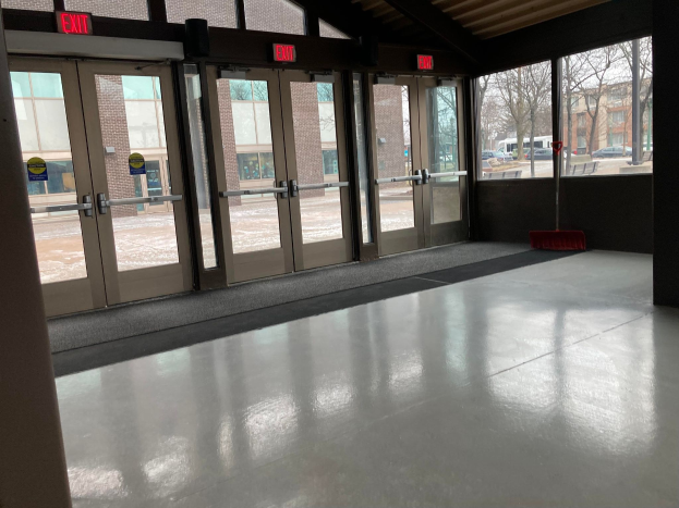 Großer Raum mit hoher Decke, mehreren Fenstern und Türen, einem Besen auf dem Boden, Schilder mit Text, ein sichtbares Dach, Bäume draußen, Fahrzeuge auf dem Boden, benachbarte Gebäude mit Fenstern und ein bewölkter Himmel, was auf einen kommerziellen Bodenreinigungsdienst hinweist.