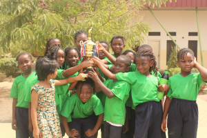 Gruppe von Kindern mit Pokal in einem Schulhof mit Bäumen und einem Gebäude im Hintergrund.