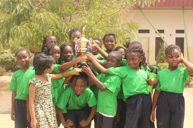 Gruppe von Kindern mit Pokal in einem Schulhof mit Bäumen und einem Gebäude im Hintergrund.