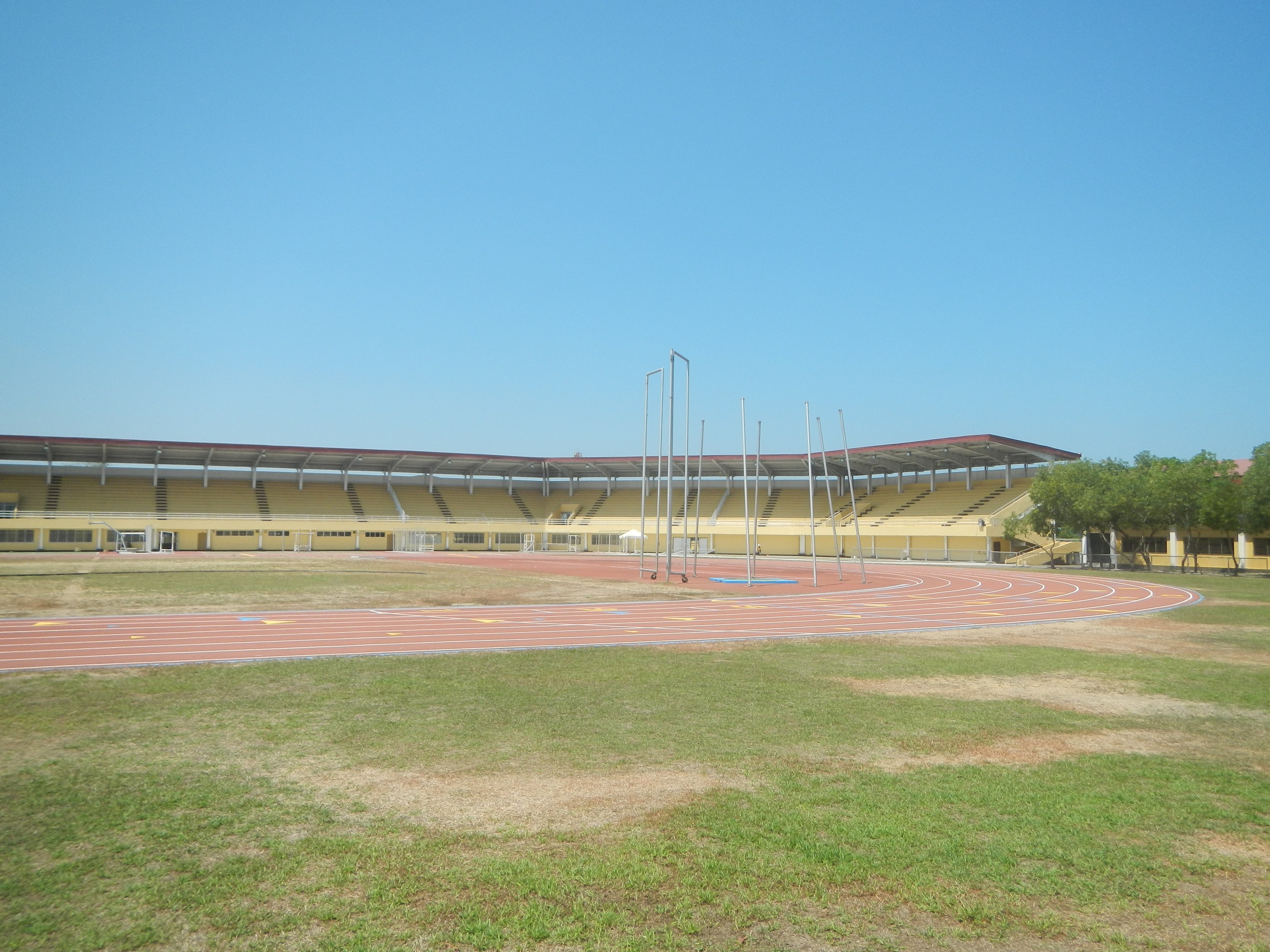 Großes Stadion mit einer zentralen Laufbahn, umgeben von grünem Gras und Bäumen auf der rechten Seite, Pfosten im Hintergrund und einem klaren blauen Himmel.