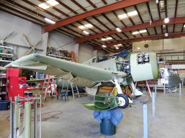 Ein großer Hangar mit verschiedenen Flugzeugen und Geräten, darunter eine Leiter, einen Stuhl, einen Tisch mit Gegenständen und Deckenleuchten.