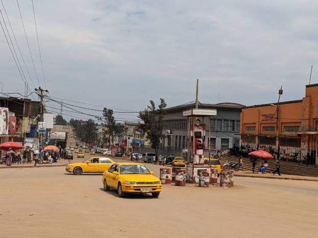 Ein gelbes Taxi fährt auf einer belebten Stadtstraße mit Gebäuden an den Seiten, während Fußgänger mit Schirmen auf dem Gehweg unter einem bewölkten Himmel gehen, mit Strommasten und Drähten im Hintergrund.
