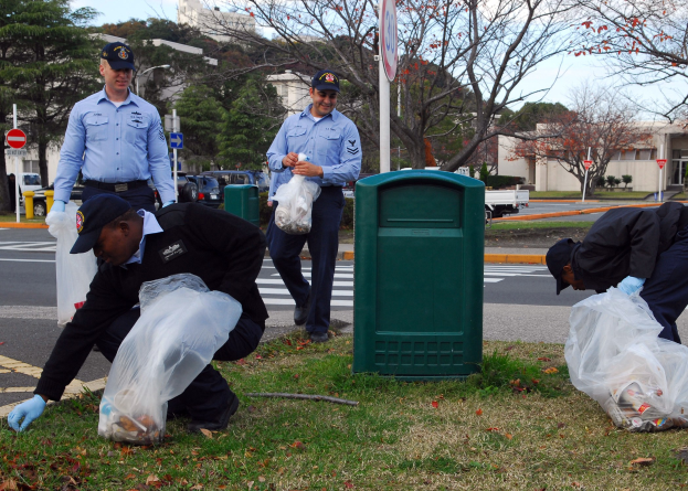 Drei Polizisten in Uniform sammeln Müll am Straßenrand, mit Bäumen, Gebäuden, Fahrzeugen und einem Mülleimer im Hintergrund.