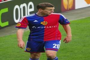 Ein Mann in einem roten und blauen Fußballtrikot läuft auf dem Feld und breitet die Arme aus, während einige Menschen auf den Tribünen sitzen und eine Tafel im Hintergrund zu sehen ist.