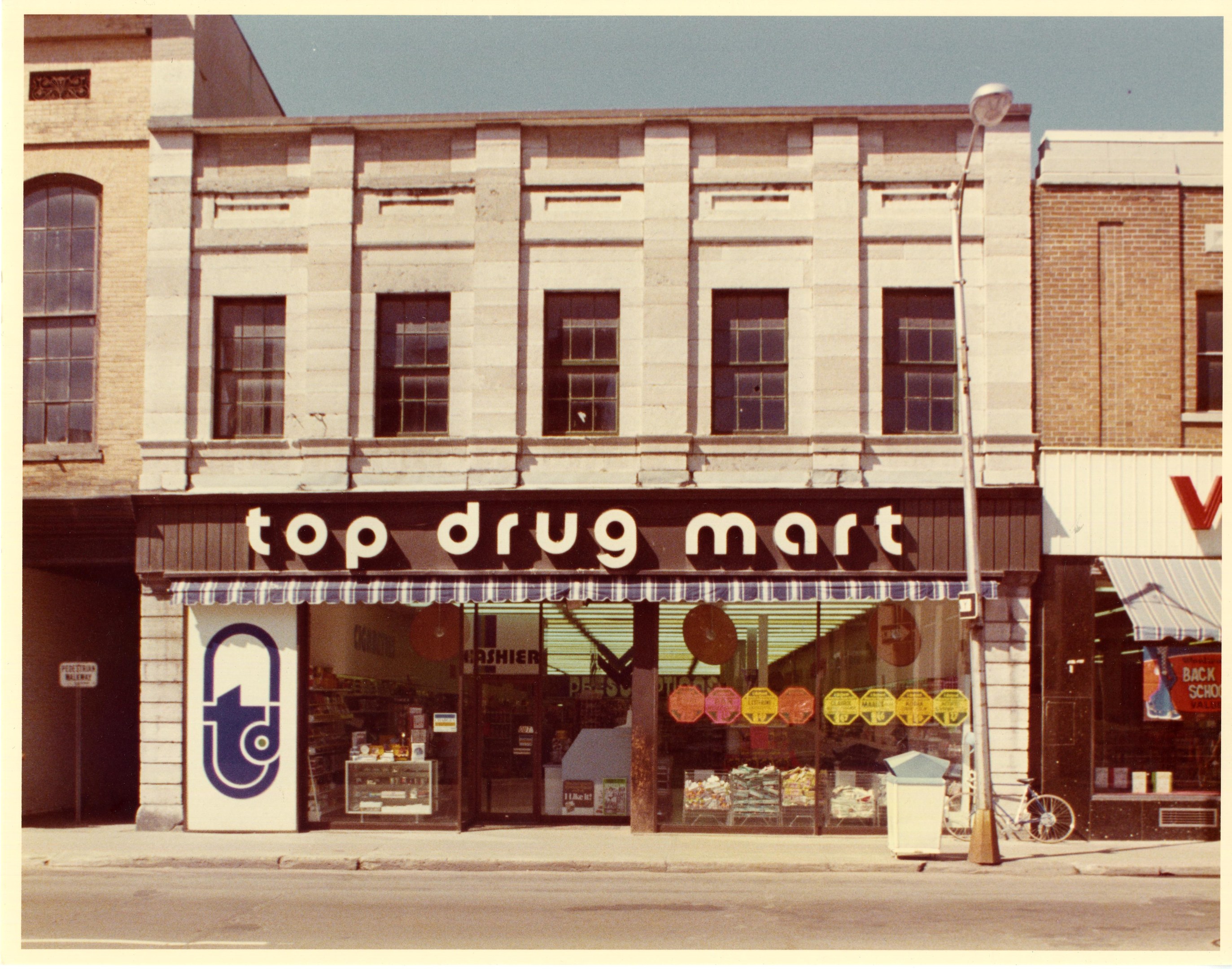 Eckladen namens "Top Drug Mart" mit Fenstern, einem Schild, einem Straßenschild, einem geparkten Fahrrad, einem Müllcontainer und einem bewölkten Himmel.