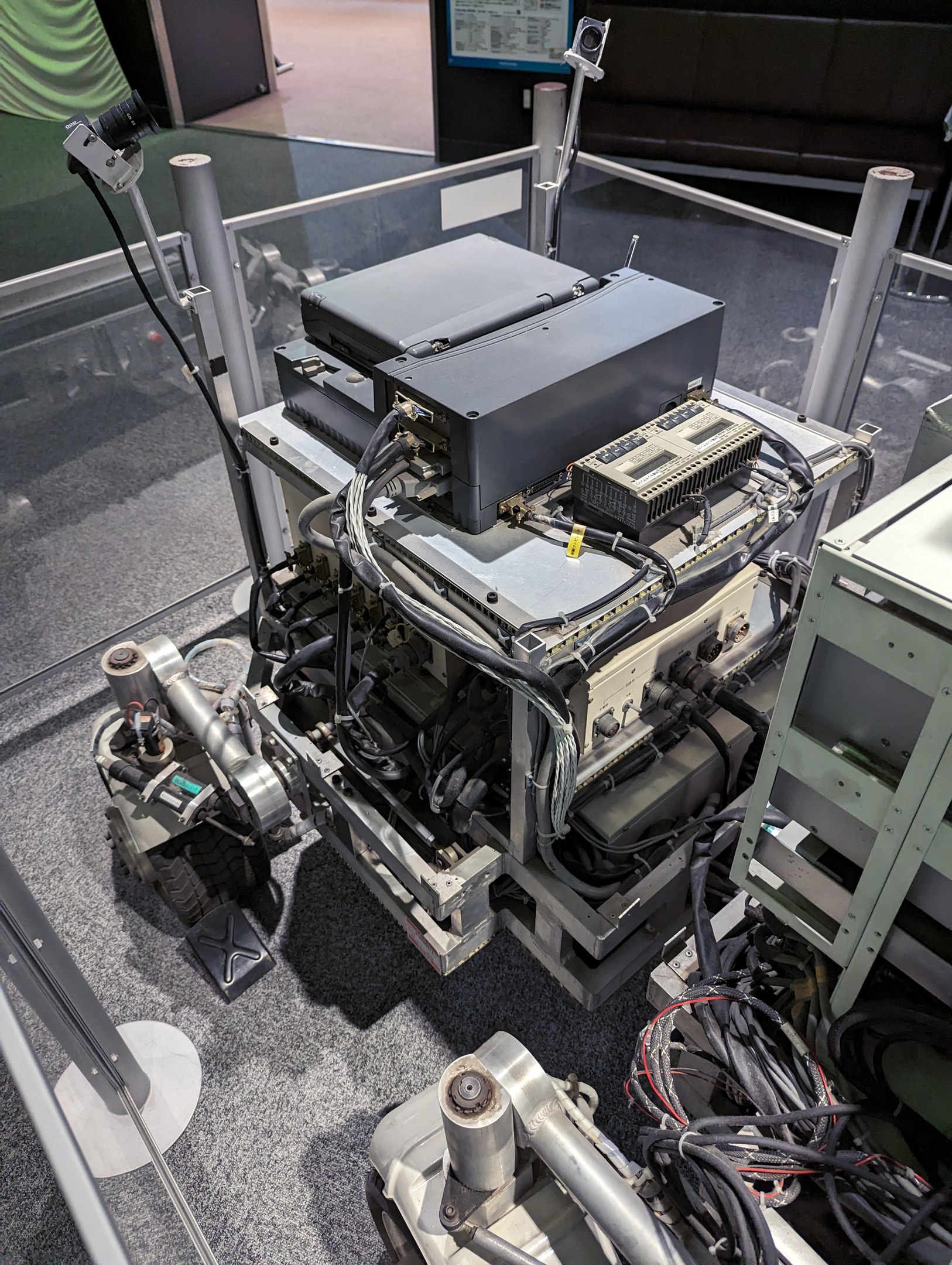 Ein Computer mit verbundenen Komponenten und Kabeln steht auf einem Tisch neben einer Glaswand, mit einer Person rechts daneben, in einem Raum mit Sofas, einem Bildschirm und sichtbarem Boden.