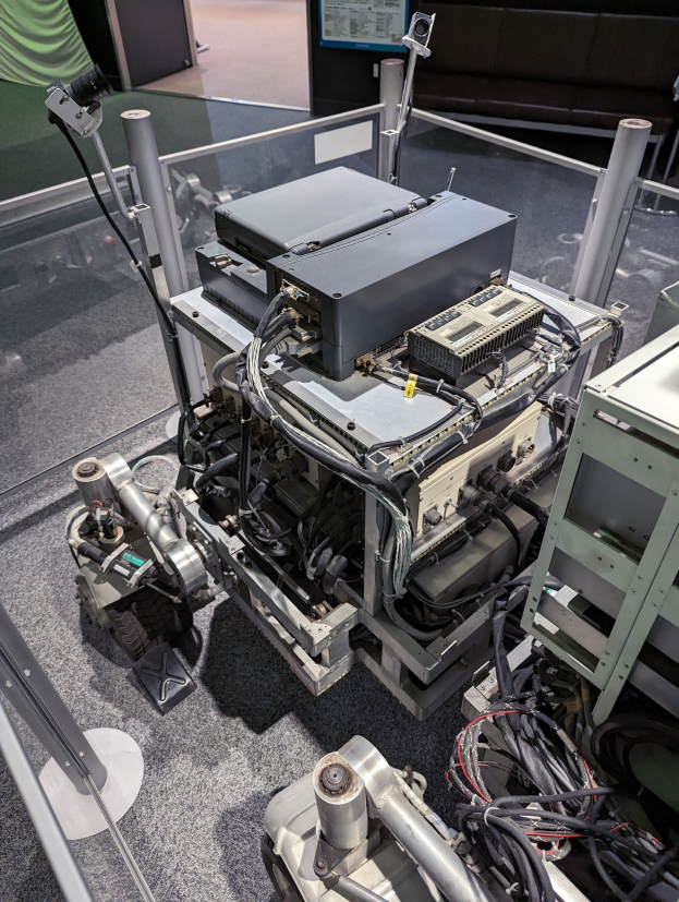 Ein Computer mit verbundenen Komponenten und Kabeln steht auf einem Tisch neben einer Glaswand, mit einer Person rechts daneben, in einem Raum mit Sofas, einem Bildschirm und sichtbarem Boden.