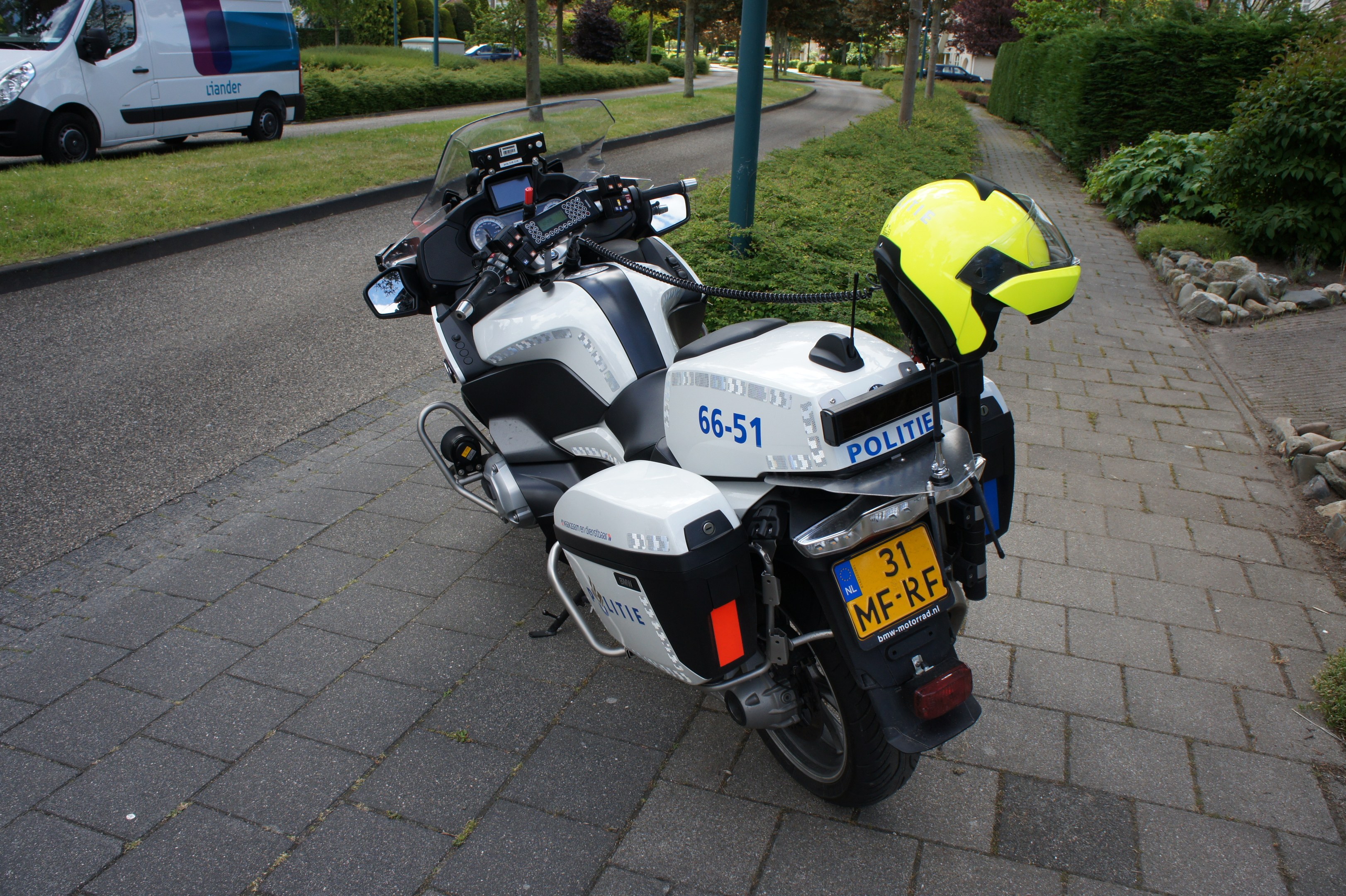 Eine Polizei-Motorrad am Straßenrand mit einem Helm darauf, umgeben von Bäumen, Pfählen, Gebäuden, Pflanzen, Gras, Steinen und anderen Fahrzeugen.