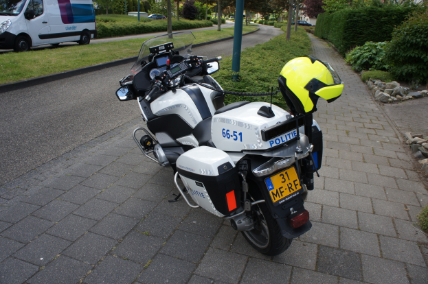 Eine Polizei-Motorrad am Straßenrand mit einem Helm darauf, umgeben von Bäumen, Pfählen, Gebäuden, Pflanzen, Gras, Steinen und anderen Fahrzeugen.