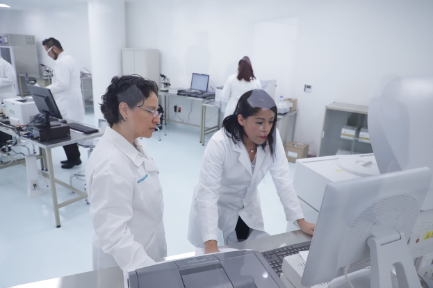 Eine Gruppe von Menschen in weißen Laborkitteln, die an Computern an Tischen in einem Labor mit Monitoren, Tastaturen, Deckenlampen, Schränken und einer Wand im Hintergrund arbeiten.