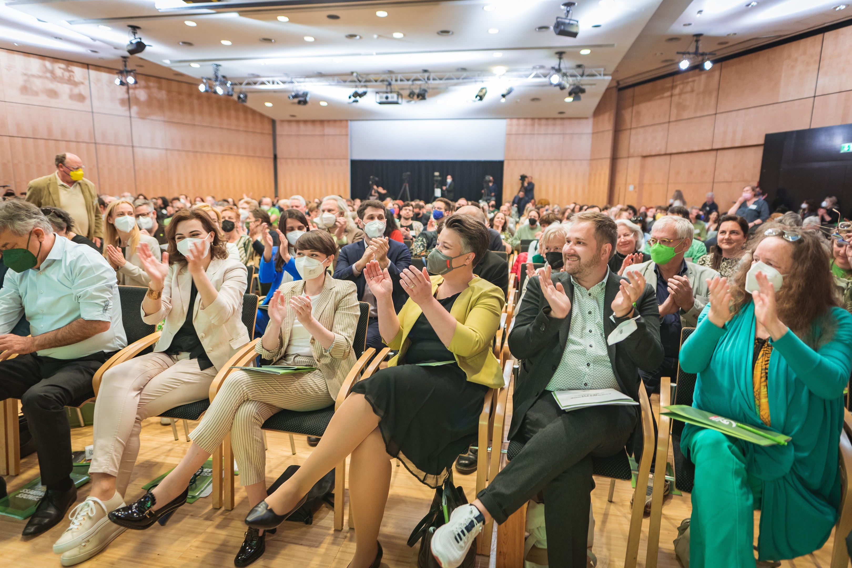 Gruppe von Menschen in Stühlen klatschen, einige tragen Masken, mit Taschen auf dem Boden, vor einem Publikum bei einer Coronavirus-Konferenz mit einem Bildschirm und Deckenleuchten im Hintergrund.