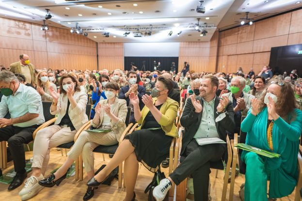 Gruppe von Menschen in Stühlen klatschen, einige tragen Masken, mit Taschen auf dem Boden, vor einem Publikum bei einer Coronavirus-Konferenz mit einem Bildschirm und Deckenleuchten im Hintergrund.